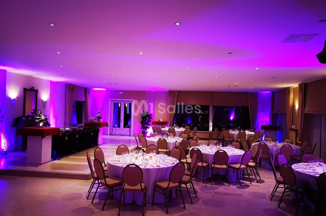 Salle de réception décorée avec des tables rondes dressées, éclairage d'ambiance violet et baies vitrées en arrière-plan.