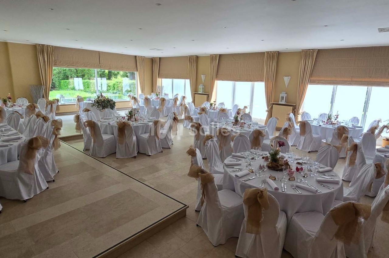 Salle de réception lumineuse avec tables rondes décorées, nappes blanches et chaises ornées de nœuds beiges.