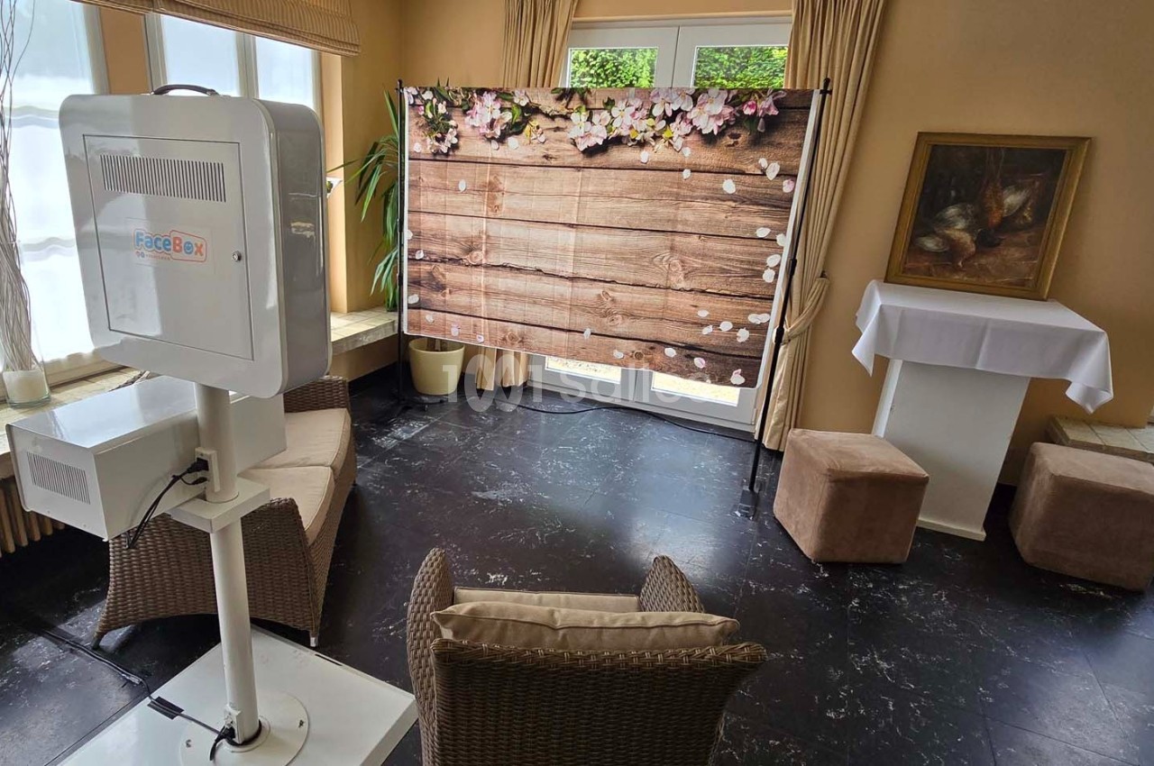 Photobooth installé dans une pièce lumineuse avec un fond en bois décoré de fleurs et des meubles en rotin.