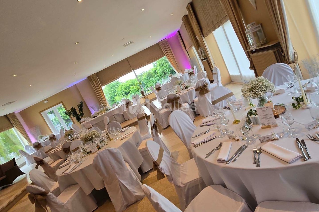 Salle de réception lumineuse avec tables rondes dressées, nappes blanches et décorations florales.