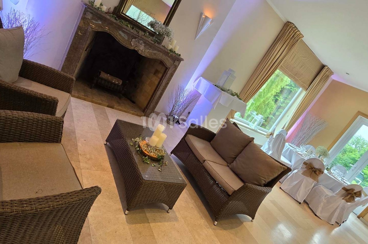 Salon lumineux avec fauteuils en rotin, cheminée décorative et vue sur une salle de réception avec grandes baies vitrées.