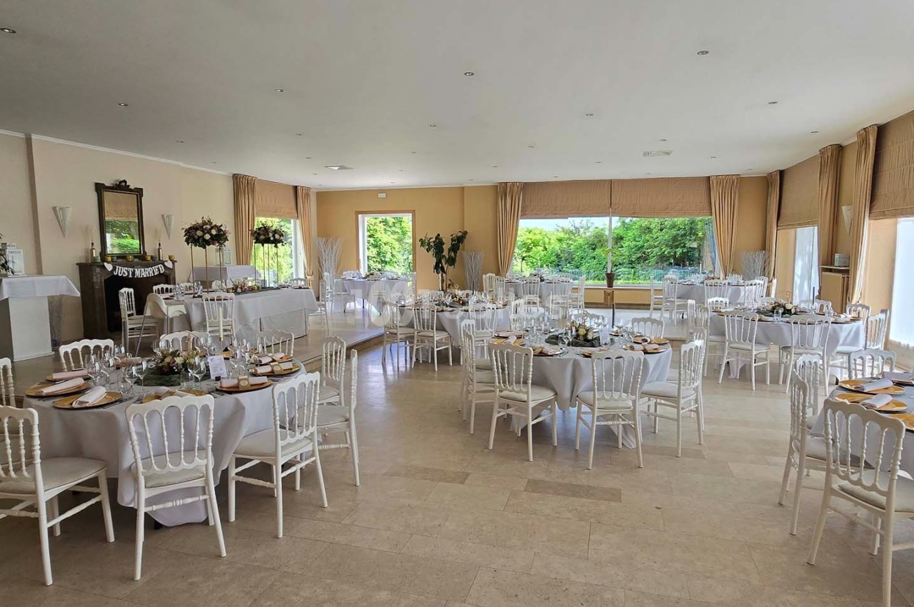 Salle lumineuse aménagée pour un événement, avec des tables rondes dressées et des chaises blanches.
