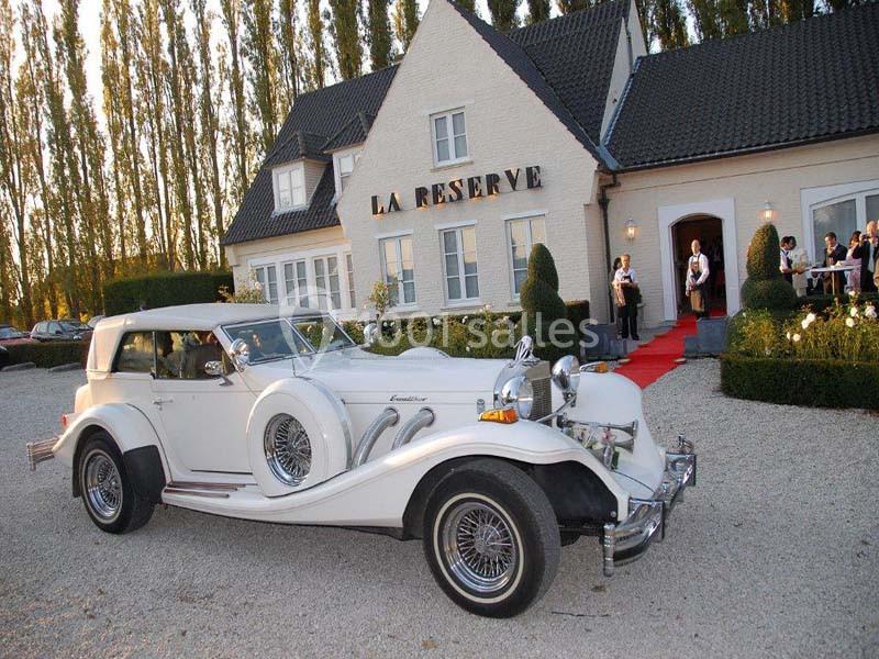 Voiture ancienne blanche garée devant un bâtiment élégant avec enseigne ’La Réserve’ et tapis rouge à l'entrée.