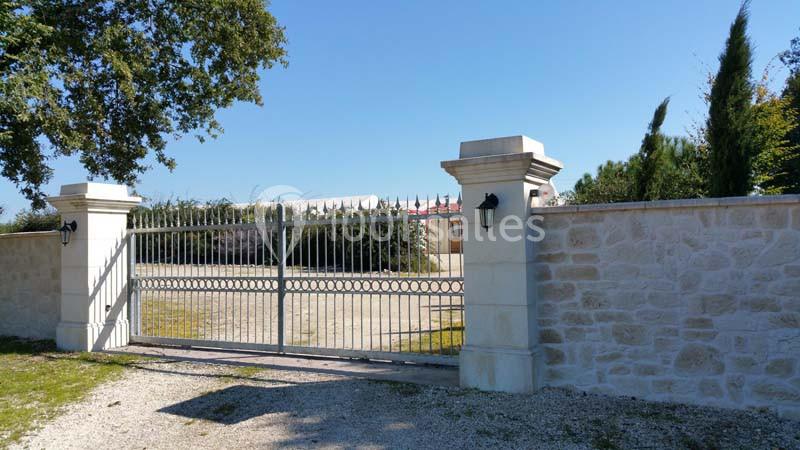 Portail métallique gris fermé entre deux piliers en pierre, entouré d'un mur et d'arbres sous un ciel bleu.