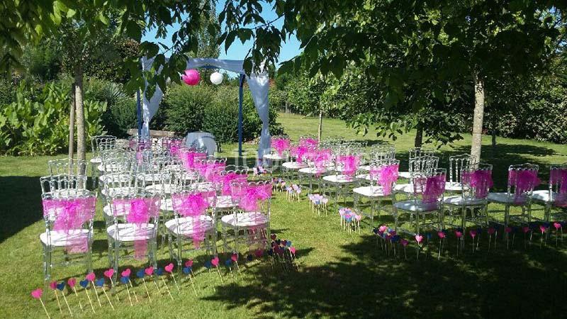 Chaises transparentes décorées de rubans roses disposées en extérieur pour une cérémonie sous des arbres.