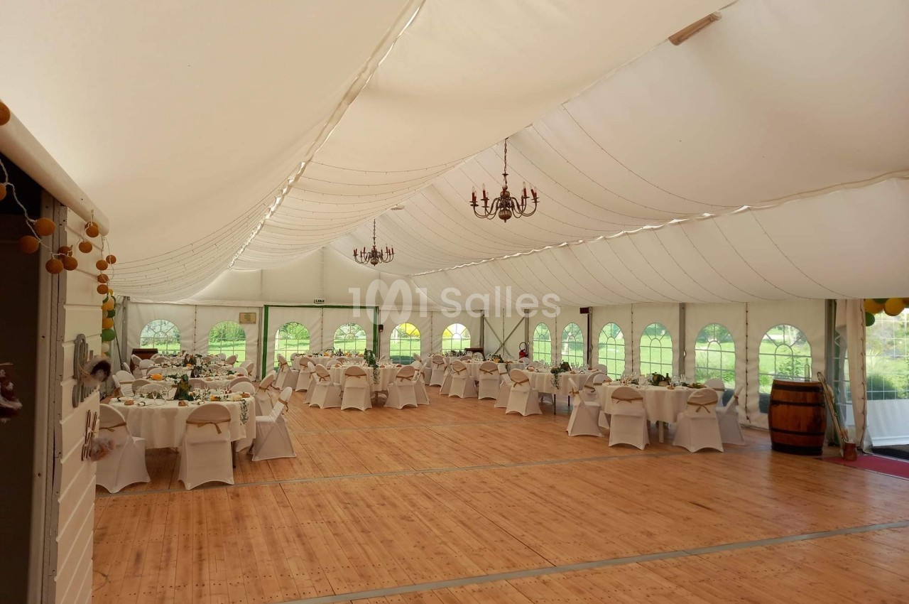 Salle de réception sous chapiteau avec tables dressées, chaises blanches et parquet en bois, éclairée par des lustres.