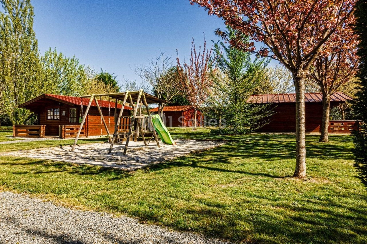 Aire de jeux avec balançoire et toboggan entourée de chalets en bois dans un espace vert arboré.
