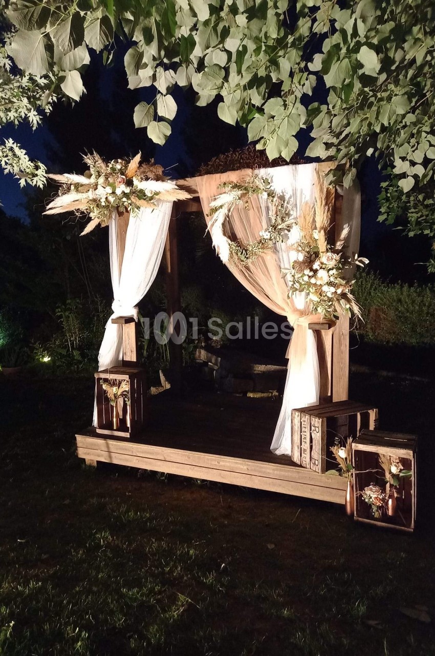 Arche de mariage en bois décorée de voilages blancs, fleurs et feuillages, éclairée dans un jardin en soirée.