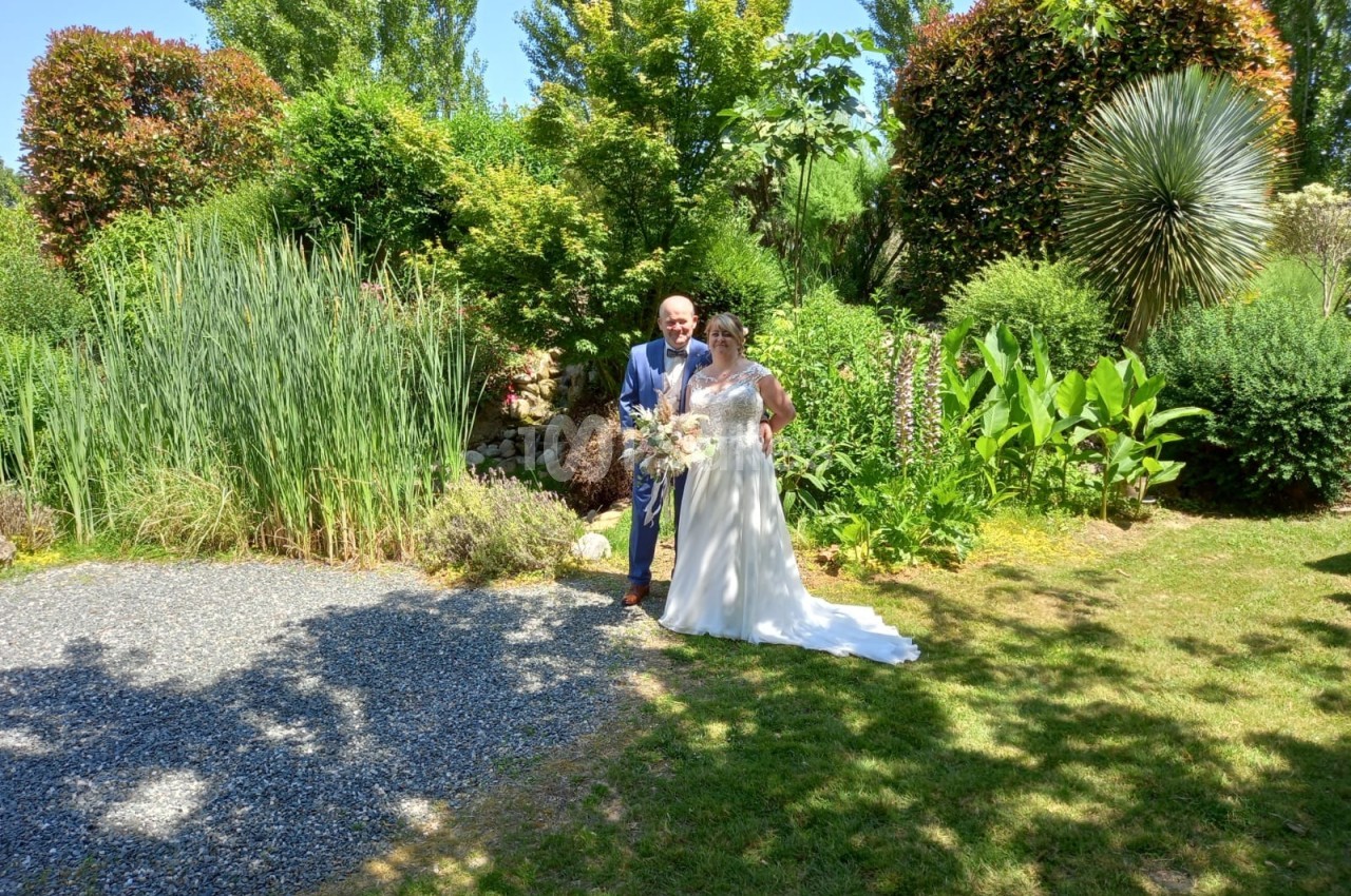 Un couple en tenue de mariage pose dans un jardin verdoyant sous un ciel ensoleillé.