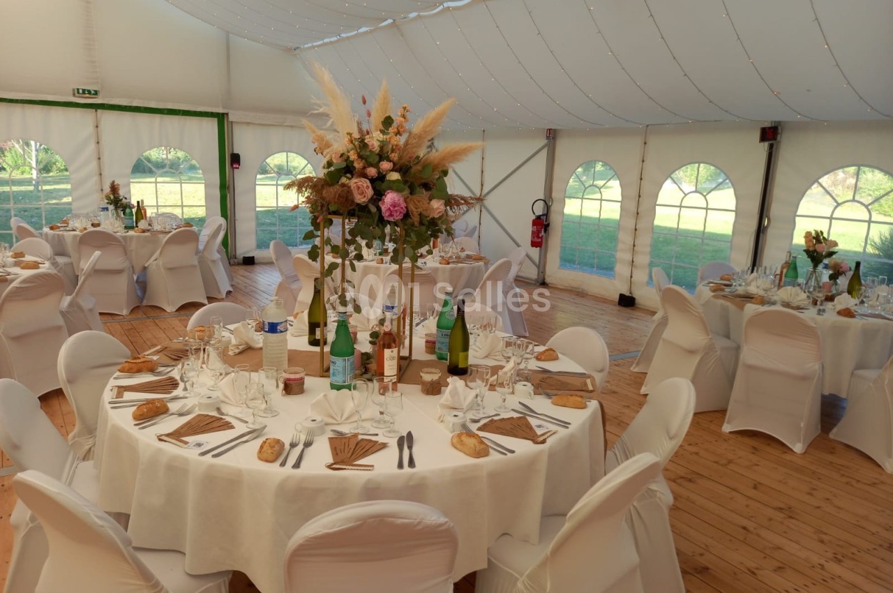 Salle de réception sous chapiteau avec tables rondes dressées, nappes blanches et décoration florale centrale.