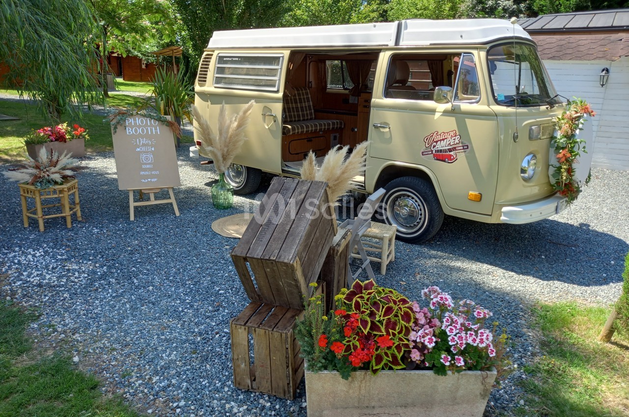 Combi vintage aménagé en photobooth, entouré de décorations florales et de caisses en bois dans un jardin.