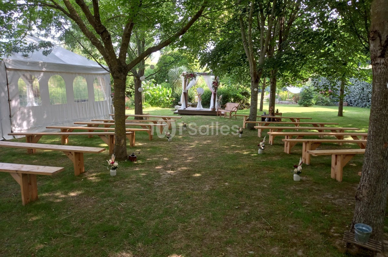 Espace extérieur aménagé pour une cérémonie, avec bancs en bois, tonnelle décorée et tente blanche sous des arbres.