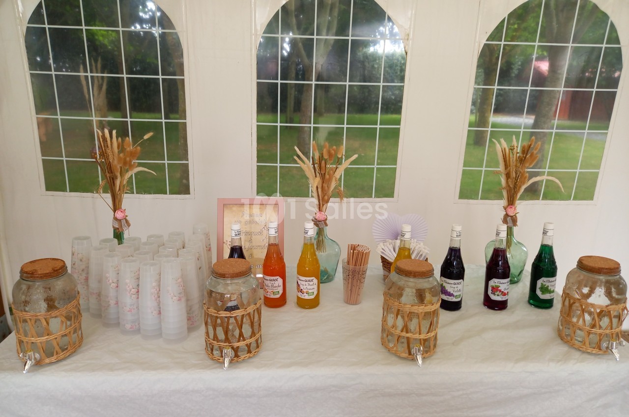 Table dressée avec des bouteilles de sirops colorés, gobelets, pailles et décorations florales sous une tente blanche.