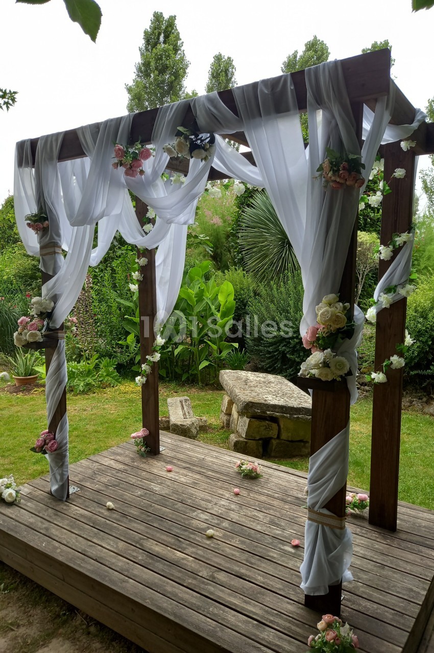 Structure en bois décorée de voilages blancs et de fleurs, installée dans un jardin verdoyant.