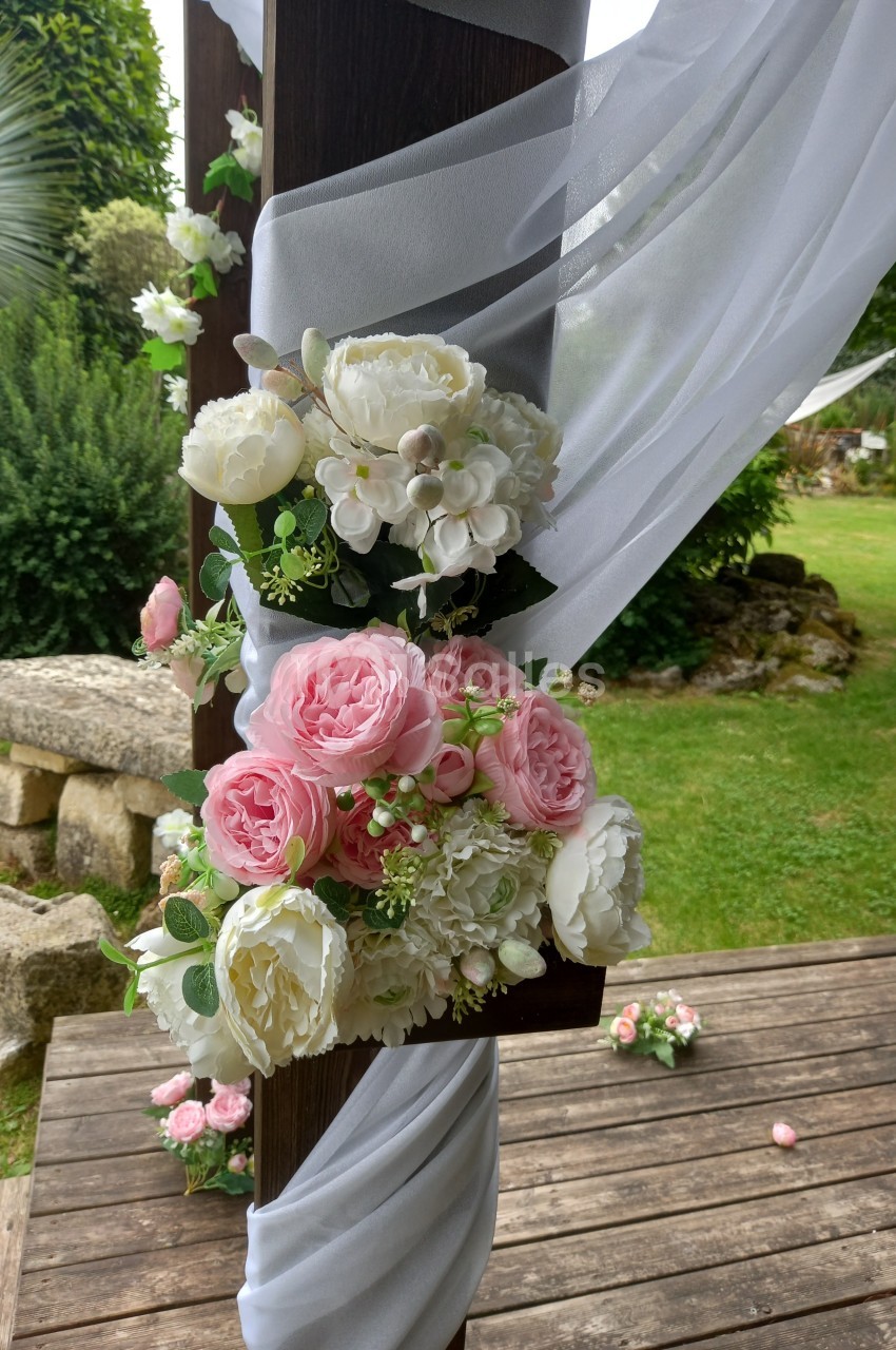 Arrangement floral avec pivoines roses et blanches attaché à une structure en bois décorée de voilages blancs, en extérieur.