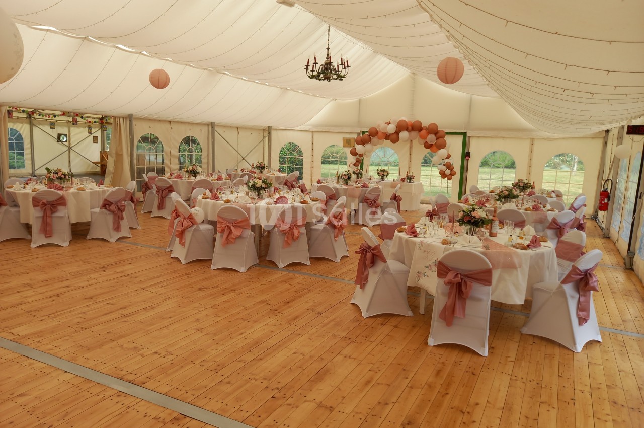 Salle de réception décorée pour un mariage, avec tables dressées, chaises ornées de nœuds roses et guirlandes lumineuses.