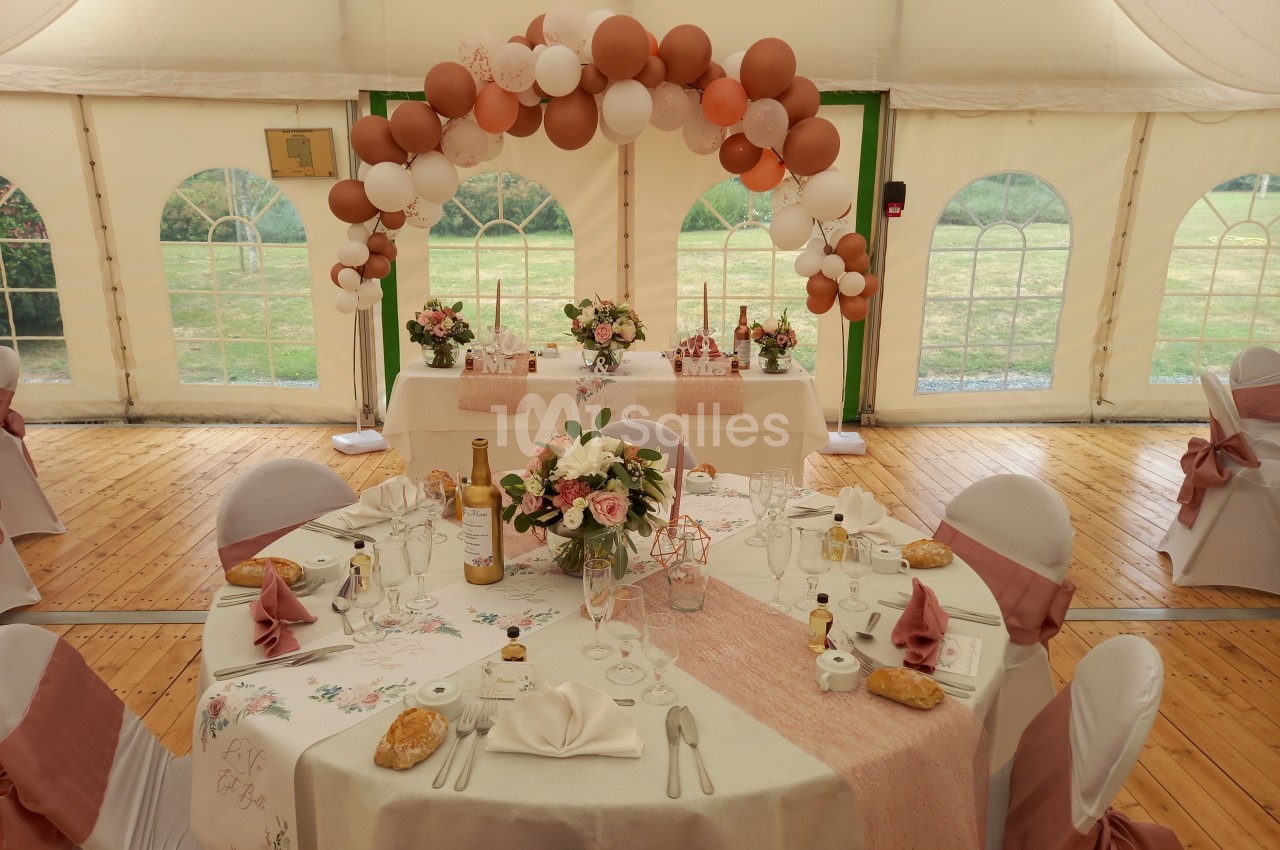 Salle de réception décorée pour un mariage avec tables dressées, nappes roses et arche de ballons dans une tente.
