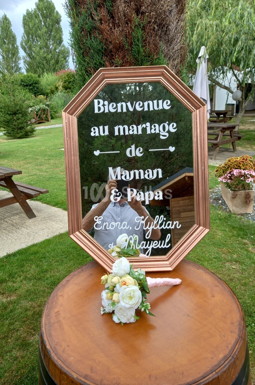 Panneau de bienvenue au mariage, posé sur un tonneau, avec des fleurs blanches, dans un jardin verdoyant.