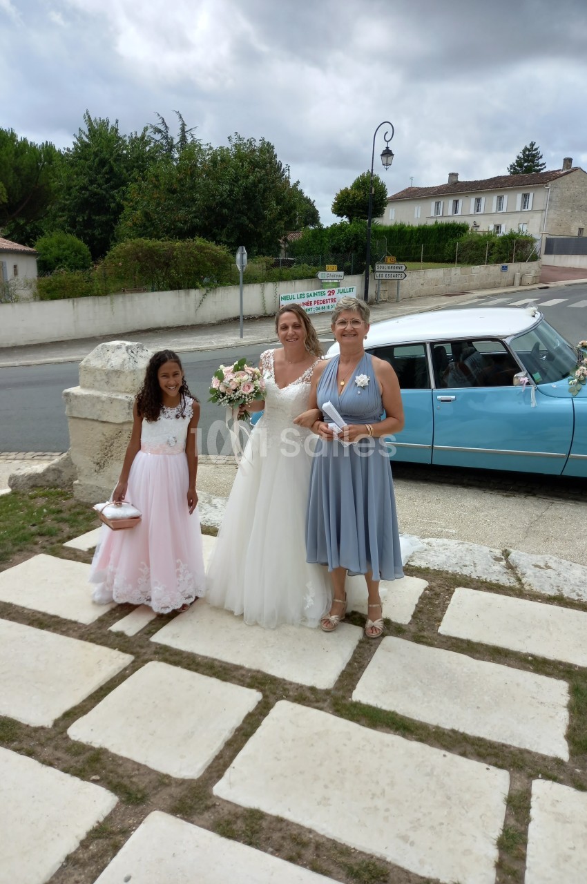 Une mariée souriante entourée de deux femmes tenant des bouquets, devant une voiture ancienne bleue dans un village.