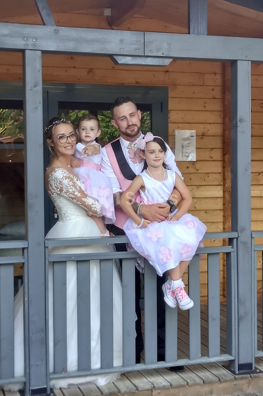 Une famille posant sur une terrasse en bois, les parents en tenue de mariage et les enfants en robes assorties.