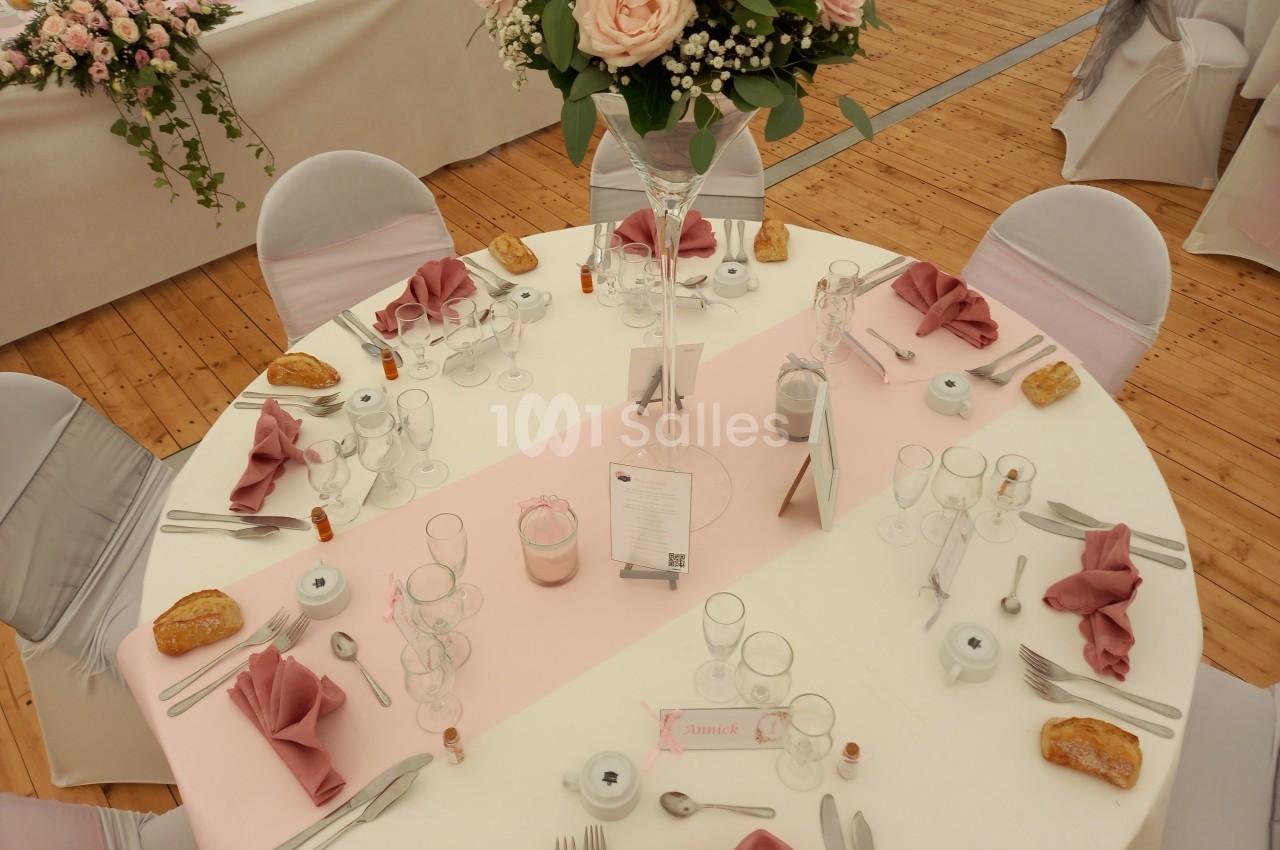 Table ronde décorée pour un mariage avec nappes blanches, serviettes roses pliées, fleurs et vaisselle disposées élégamment.