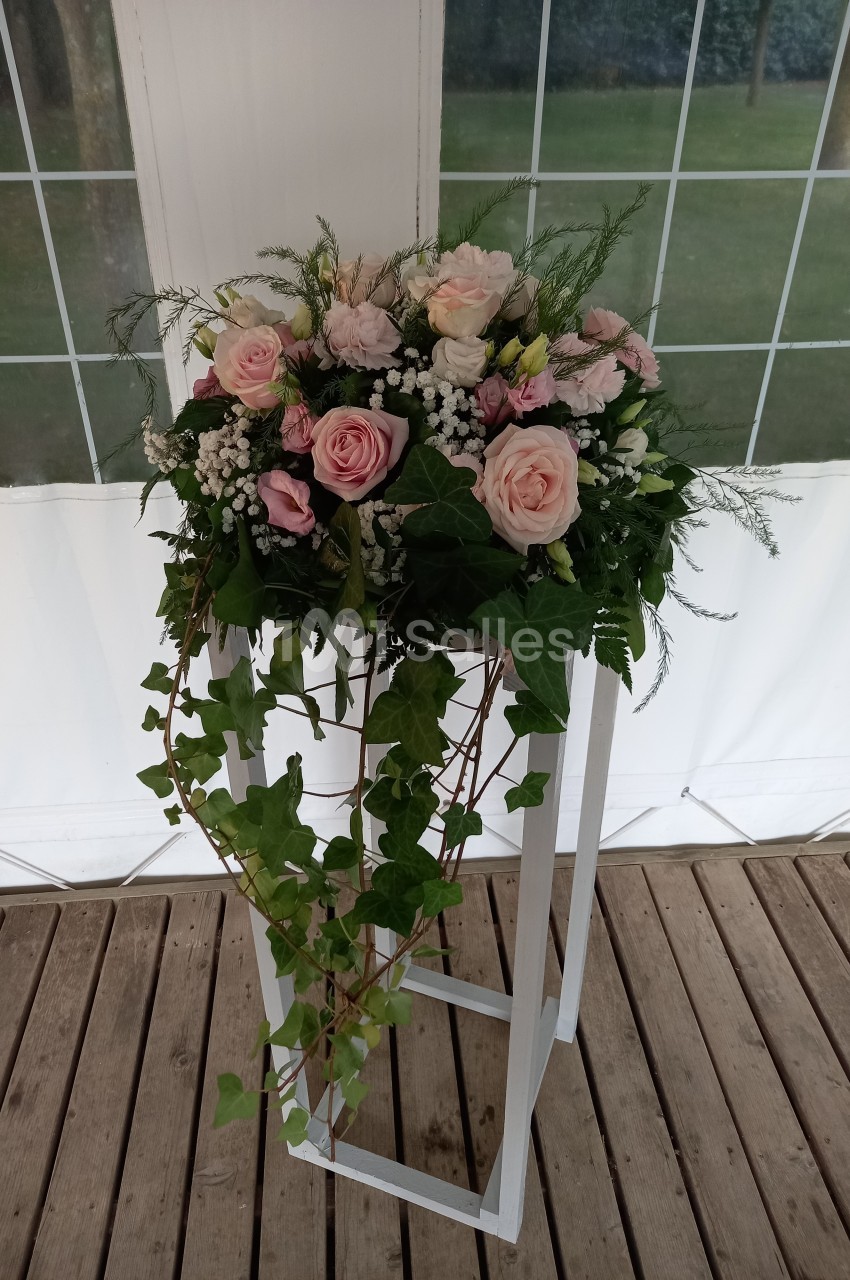 Arrangement floral avec des roses roses, du gypsophile et du lierre, posé sur un support blanc en bois sur une terrasse.