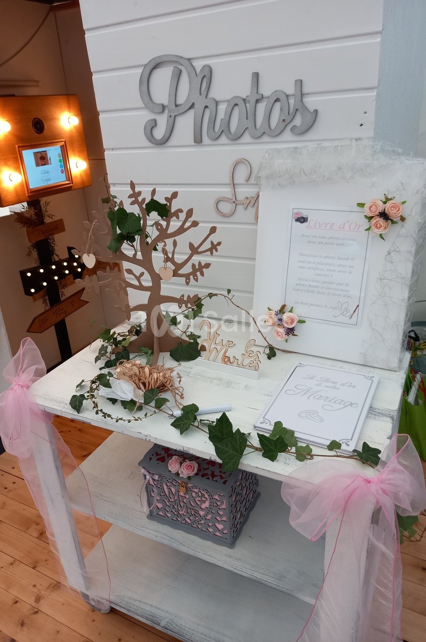 Décoration de mariage avec un arbre à vœux, des roses, des cadres et une boîte sur une table blanche ornée de rubans roses.