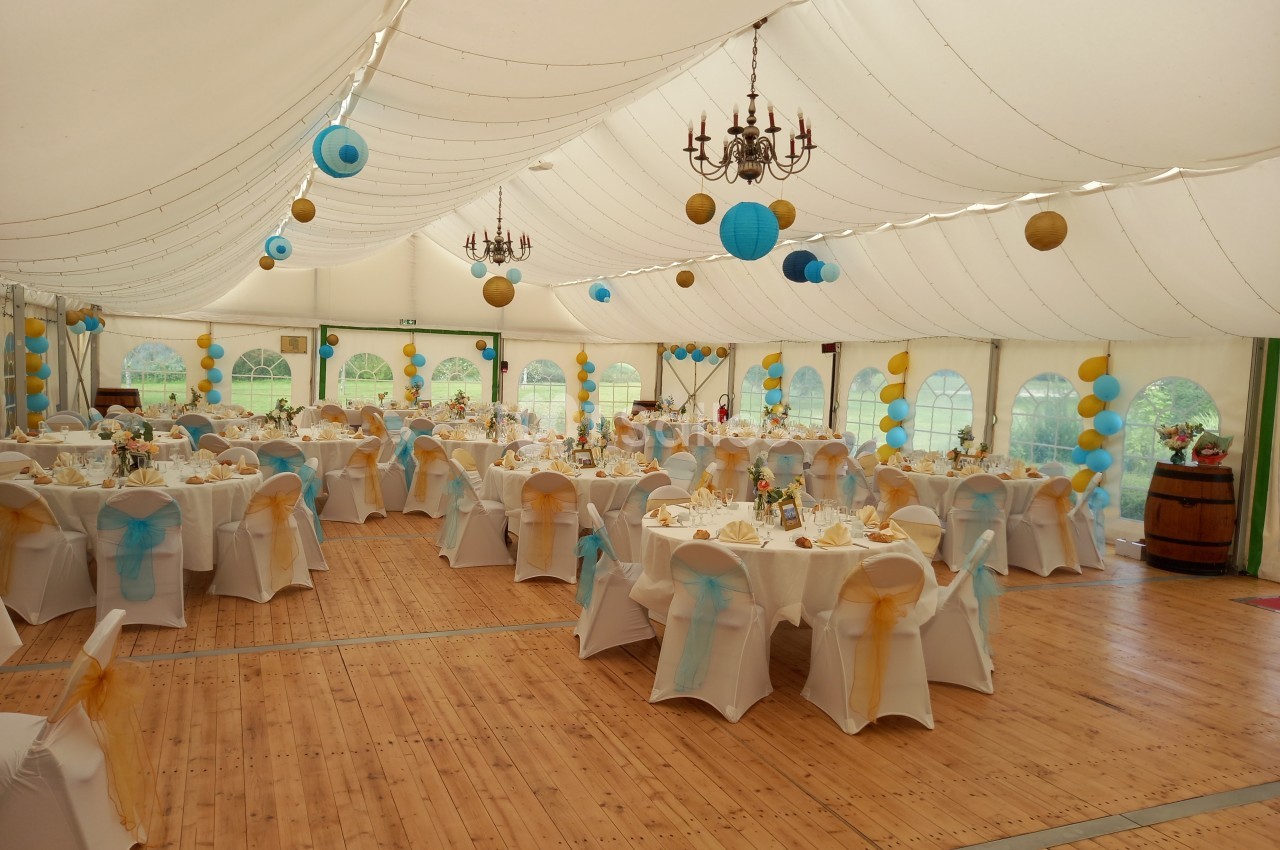 Salle de réception sous chapiteau avec tables décorées en blanc, bleu et jaune, éclairée par des lustres et des guirlandes…