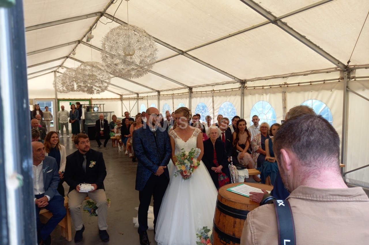 Un couple de mariés debout devant un officiant dans une tente, entourés d'invités assis.