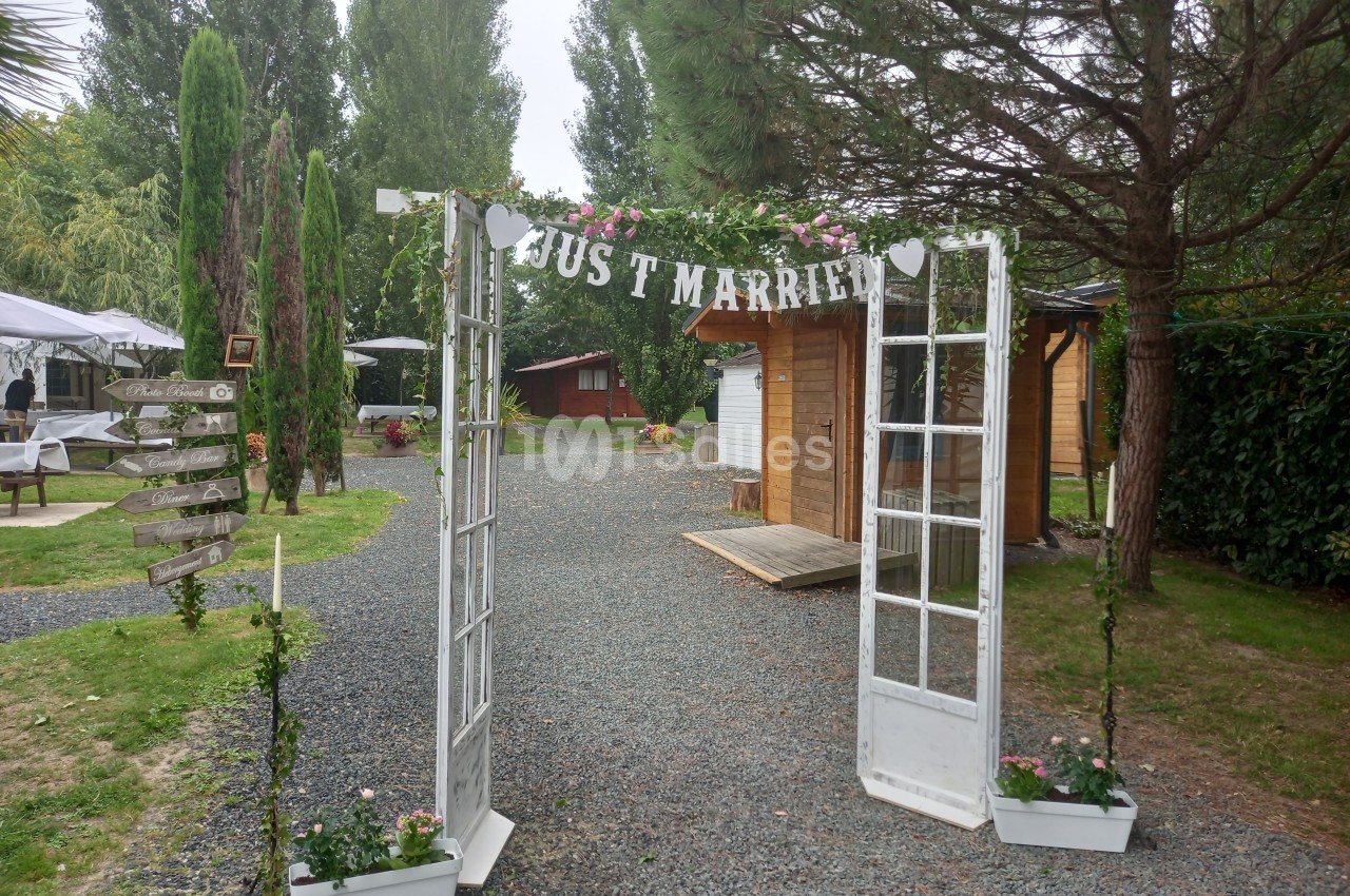 Arche décorée de fleurs et d'une bannière ’Just Married’ dans un jardin avec des cabanes en bois en arrière-plan.