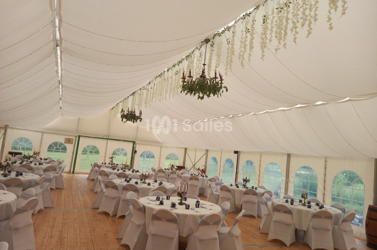 Intérieur d'une tente de réception avec tables rondes dressées, chaises blanches et décorations suspendues.