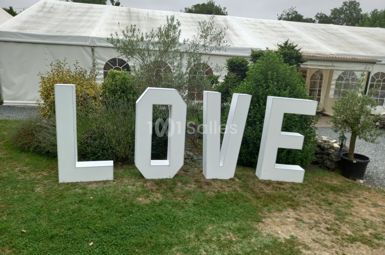 Lettres blanches formant le mot ’LOVE’ disposées sur une pelouse devant une grande tente blanche.