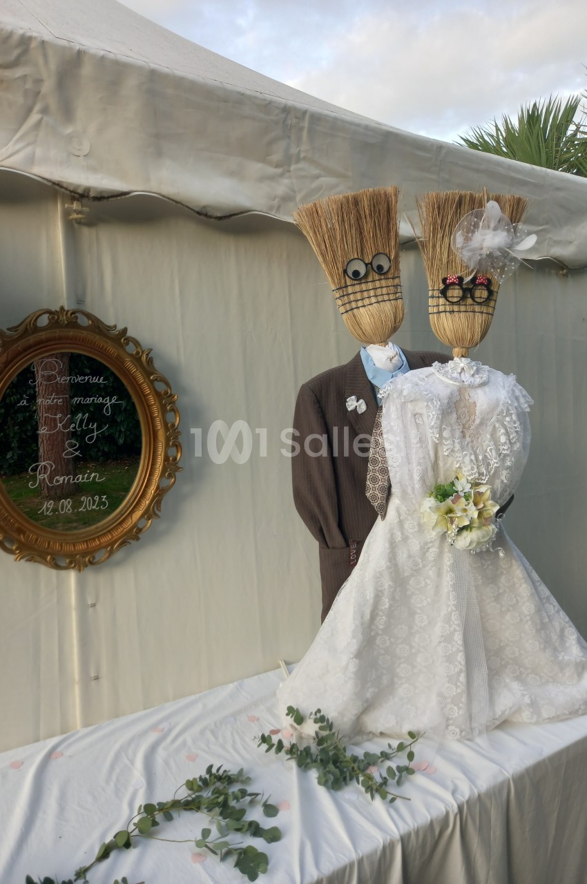 Deux balais déguisés en mariés avec robe et costume, posés devant une tente blanche et un cadre décoratif.