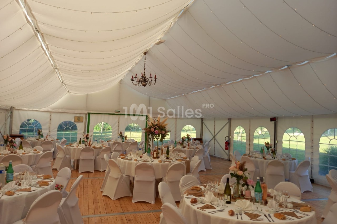 Intérieur d'une tente de réception décorée avec des tables rondes dressées, des chaises blanches et des fleurs.