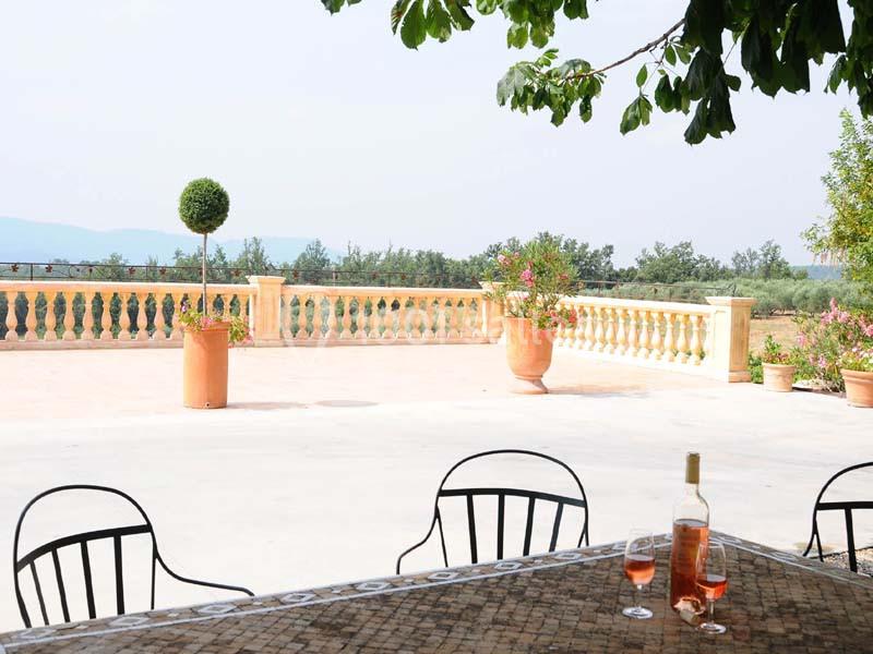 Terrasse ensoleillée avec table, chaises, balustrade, plantes en pot et vue sur la campagne.