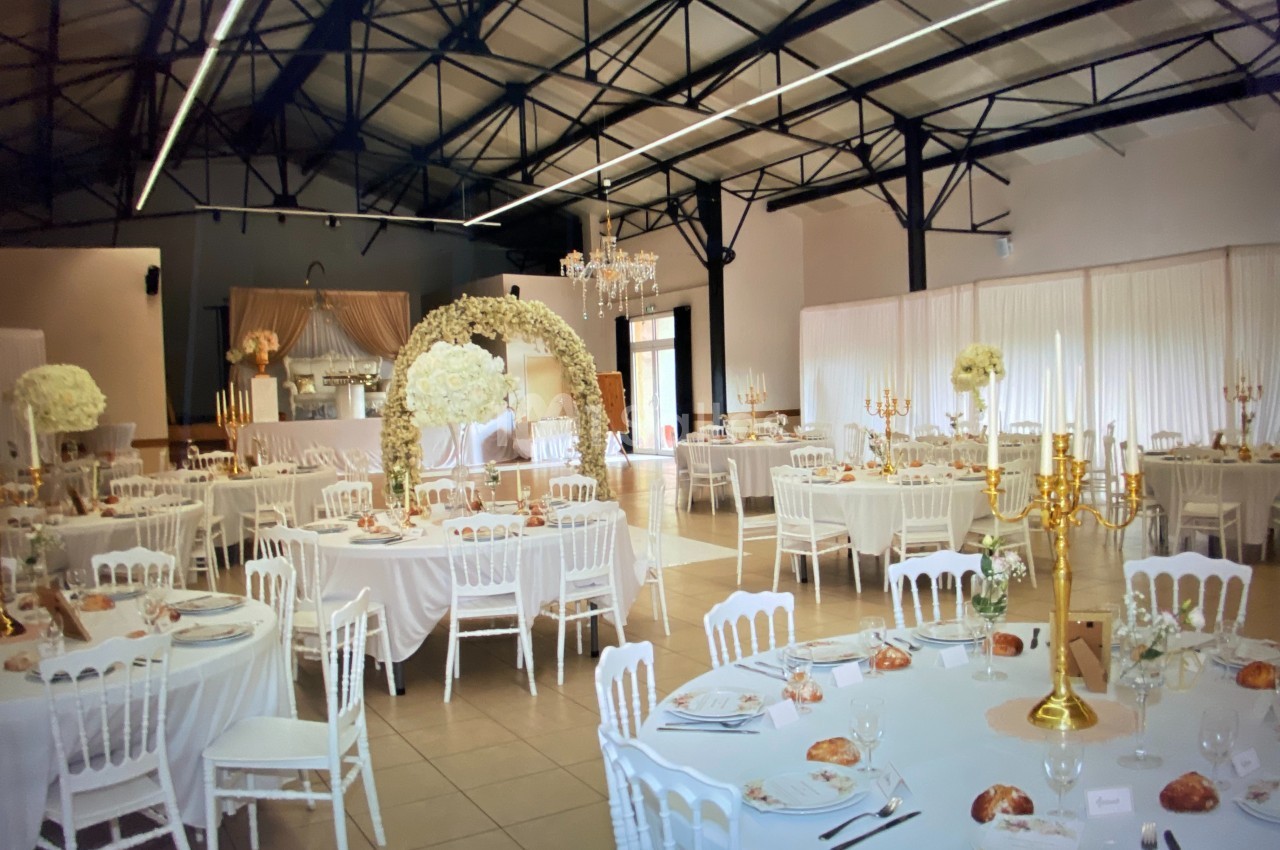 Salle de réception décorée pour un mariage avec tables rondes, nappes blanches, arche florale et chandeliers dorés.