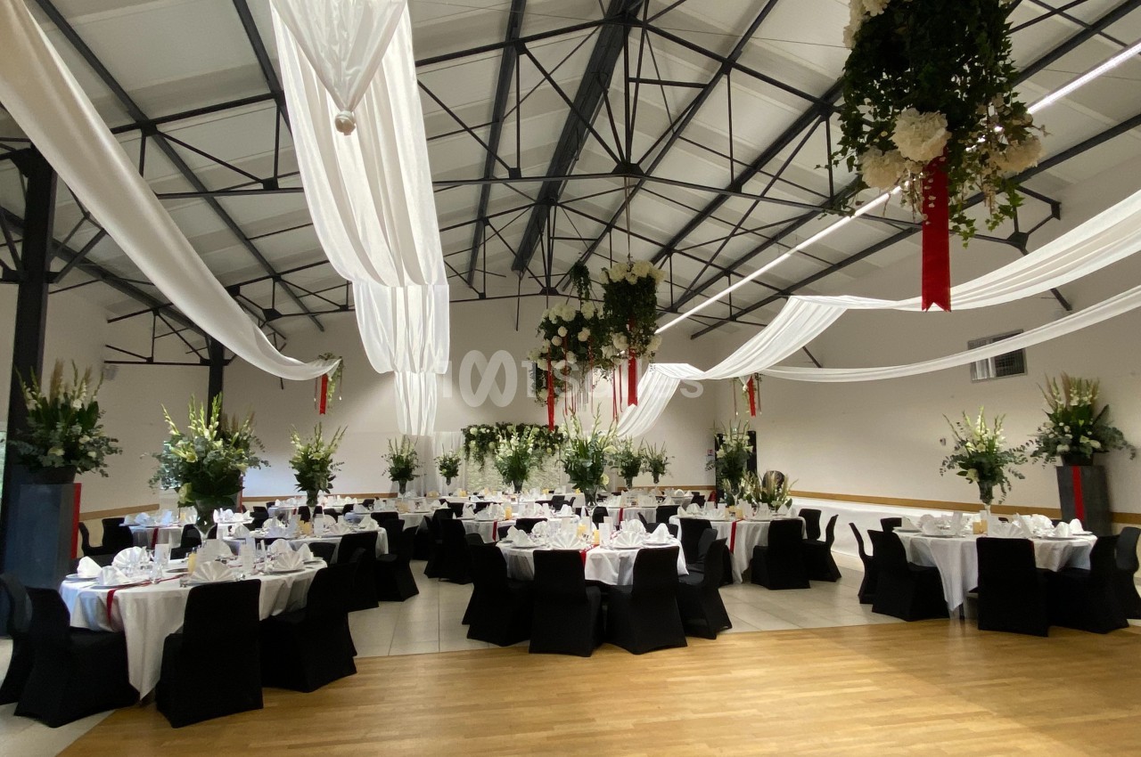 Salle de réception décorée avec des tables rondes dressées, nappes blanches, chaises noires et drapés suspendus.