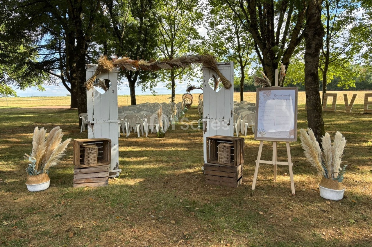 Allée de cérémonie en plein air décorée de portes blanches, pampas et chaises alignées sous des arbres.