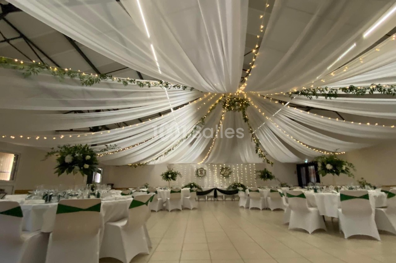Salle de réception décorée avec des drapés blancs suspendus, guirlandes lumineuses et tables élégamment dressées.