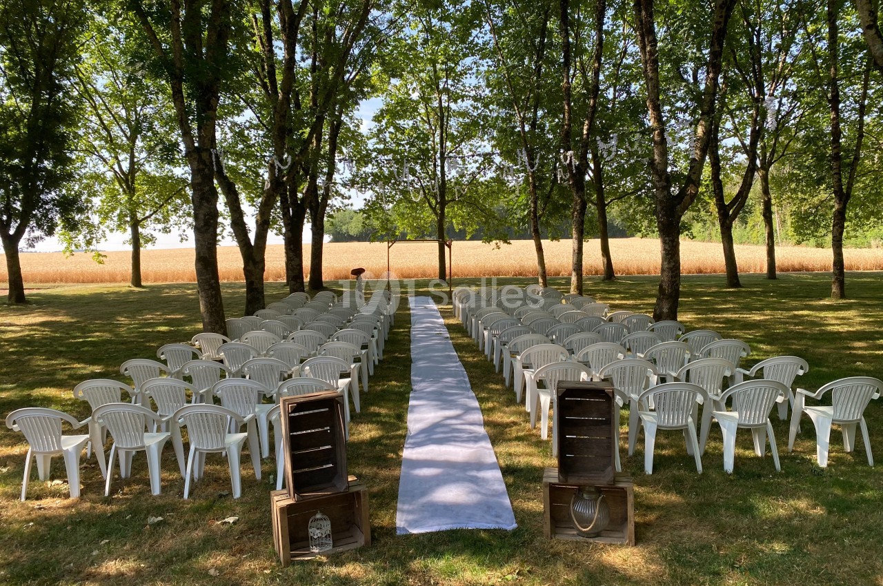 Allée centrale bordée de chaises blanches en plastique, installées dans un sous-bois pour une cérémonie en plein air.