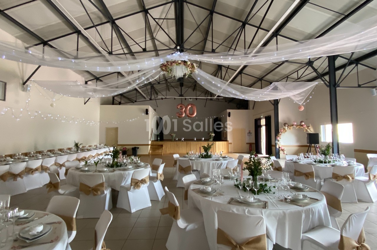 Salle décorée pour une fête avec des tables rondes dressées, des chaises ornées de nœuds et un chiffre ’30’ suspendu.