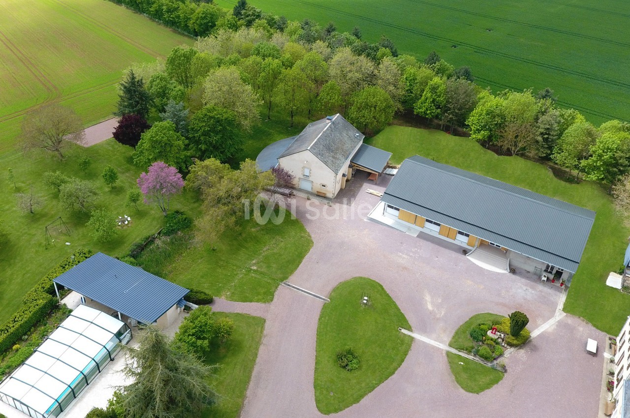 Vue aérienne d'une propriété avec maison, dépendances, piscine couverte et espaces verts entourés de champs.