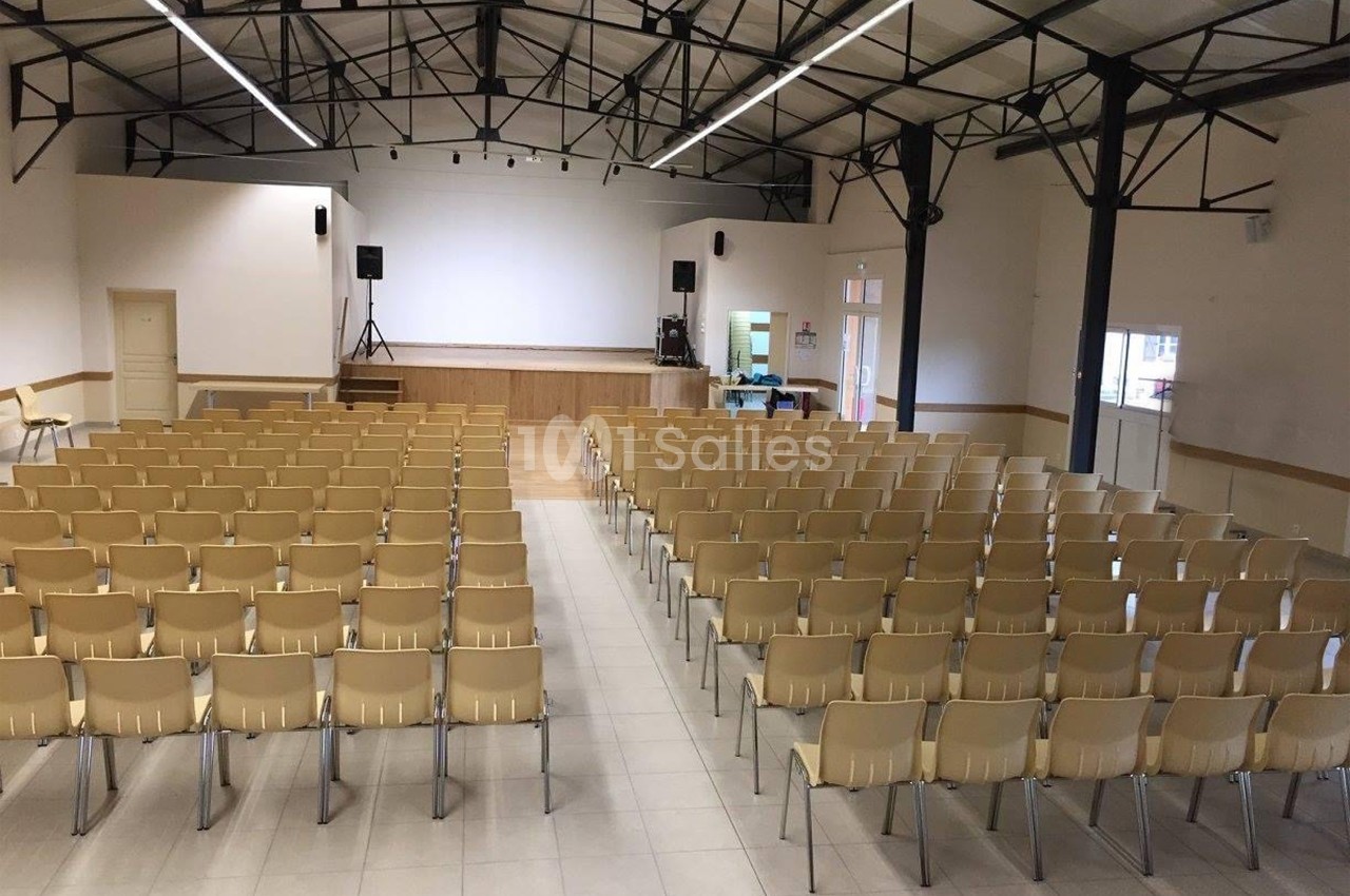 Salle avec des rangées de chaises en plastique beige face à une scène surélevée dans un espace lumineux.