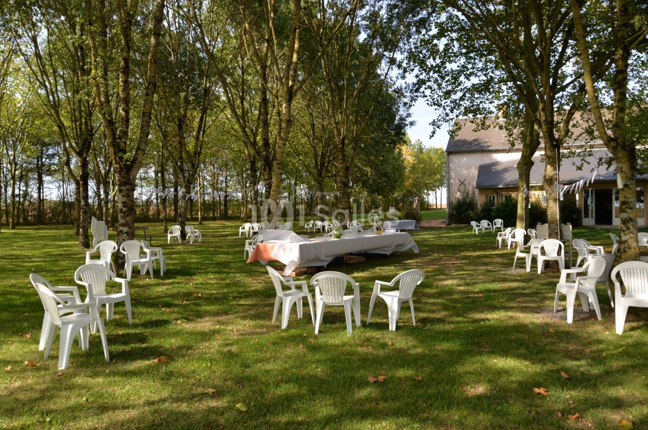 Chaises et tables blanches disposées dans un jardin ombragé près d'une maison, sous des arbres feuillus.