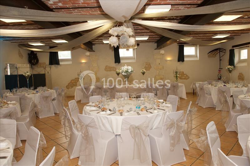 Salle de réception décorée avec des tables rondes, nappes blanches, chaises ornées de nœuds et plafonds avec drapés.