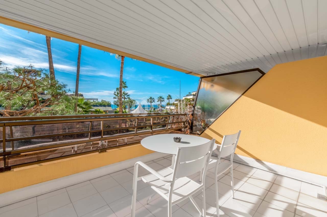 Balcon avec table et chaises blanches, vue sur des palmiers, la mer et un ciel bleu, sous un toit en panneaux blancs.