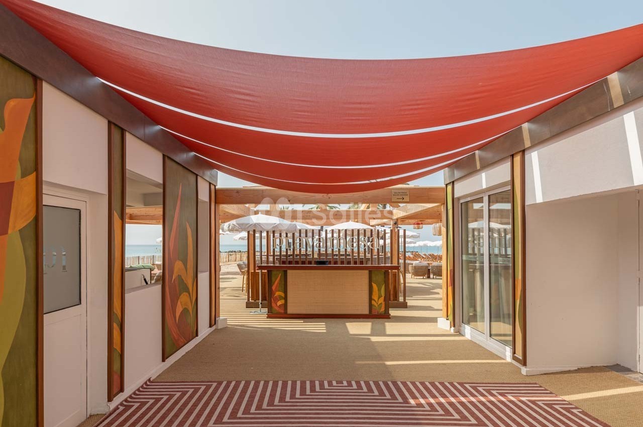 Vue d'une terrasse en bord de mer avec un toit en toile rouge, un bar central et des parasols blancs.