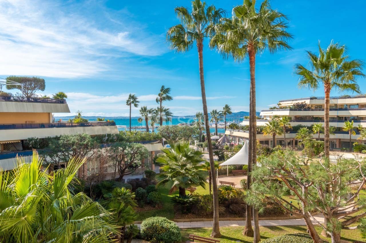Vue sur un jardin arboré avec des palmiers, des bâtiments modernes et la mer en arrière-plan sous un ciel bleu.