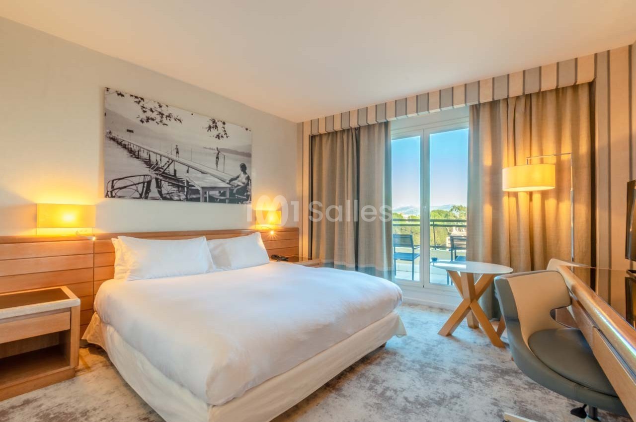 Chambre d'hôtel lumineuse avec lit double, bureau, fauteuil, grandes fenêtres et vue sur un balcon.