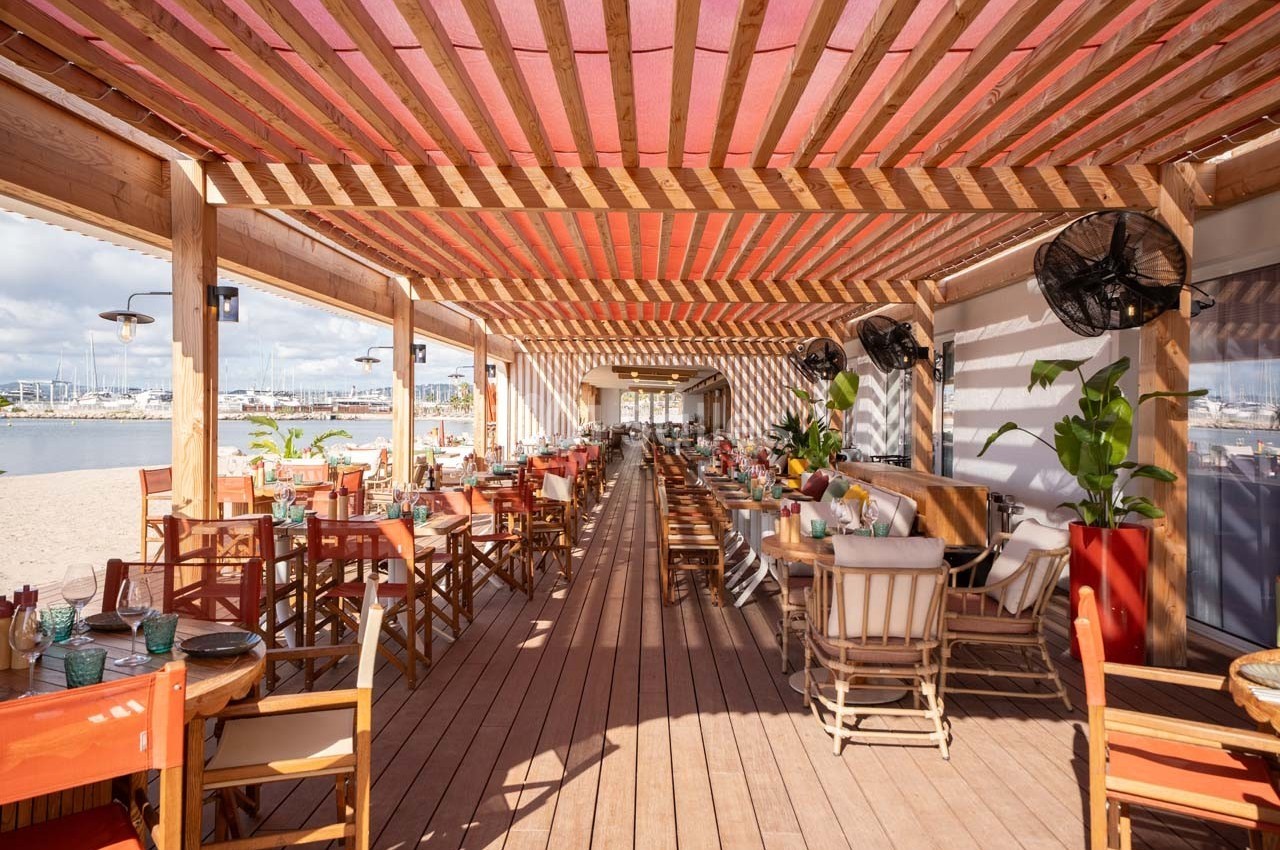 Terrasse en bois d'un restaurant en bord de plage, avec tables et chaises disposées sous une pergola.